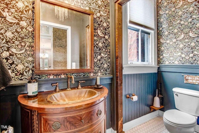 bathroom with a wainscoted wall, toilet, vanity, and wallpapered walls