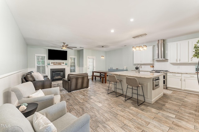 kitchen with a center island with sink, a ceiling fan, open floor plan, appliances with stainless steel finishes, and wall chimney exhaust hood