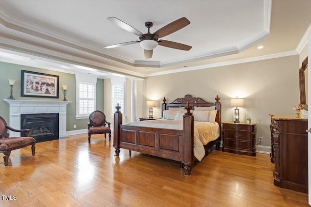 bedroom featuring a glass covered fireplace, light wood-type flooring, a raised ceiling, and baseboards