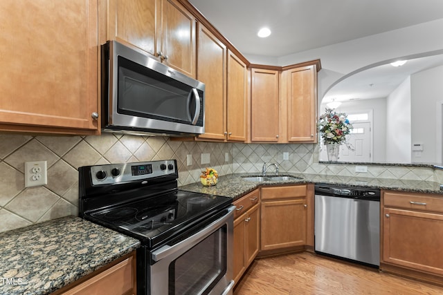 kitchen featuring light wood finished floors, dark stone counters, a sink, appliances with stainless steel finishes, and backsplash