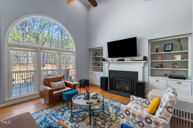 living room featuring a fireplace with flush hearth, a ceiling fan, built in features, wood finished floors, and a high ceiling