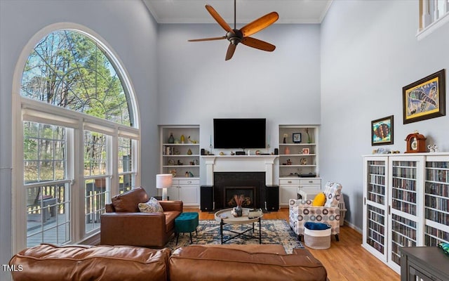 living area featuring a fireplace with flush hearth, ornamental molding, a towering ceiling, wood finished floors, and a ceiling fan