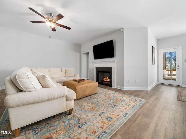 living room with a warm lit fireplace, baseboards, a ceiling fan, and wood finished floors