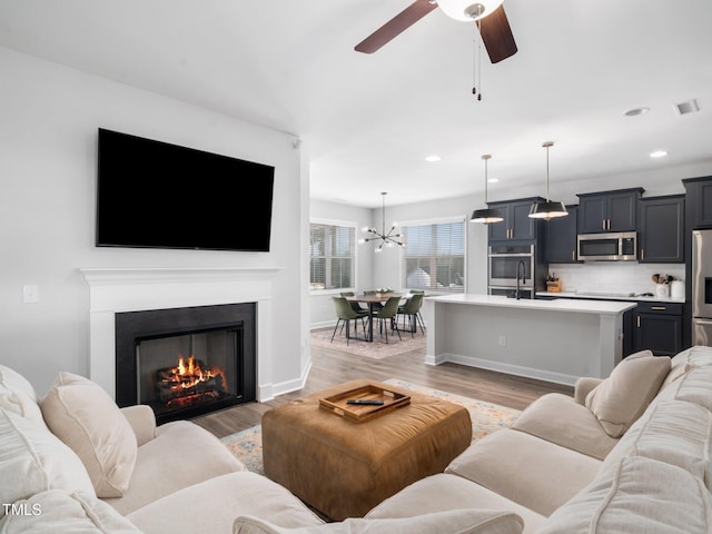 living room with light wood-type flooring, visible vents, ceiling fan with notable chandelier, recessed lighting, and a lit fireplace