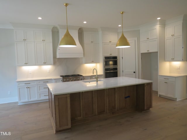 kitchen with light wood-type flooring, custom range hood, a large island with sink, white cabinetry, and a sink