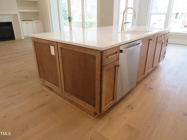 kitchen with brown cabinetry, an island with sink, a sink, light wood-style floors, and dishwasher