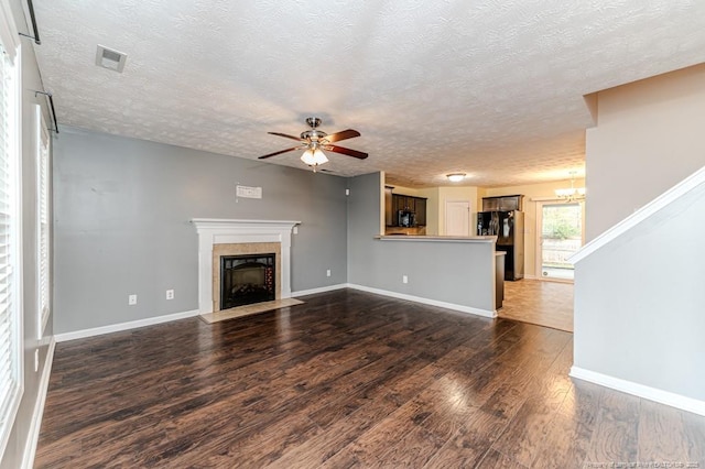 unfurnished living room featuring a fireplace with flush hearth, ceiling fan with notable chandelier, a textured ceiling, baseboards, and dark wood-style flooring