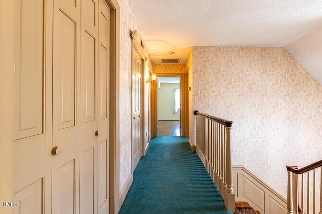 hallway featuring visible vents, an upstairs landing, a textured ceiling, carpet, and wallpapered walls