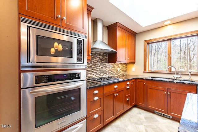 kitchen featuring decorative backsplash, dark stone countertops, stainless steel appliances, wall chimney exhaust hood, and a sink