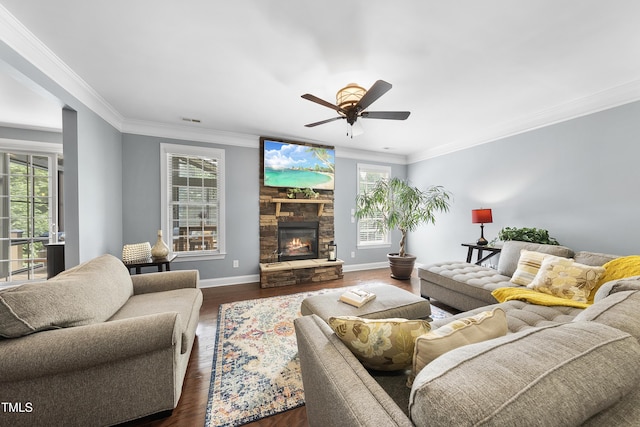 living area featuring dark wood-style flooring, crown molding, baseboards, a ceiling fan, and a stone fireplace