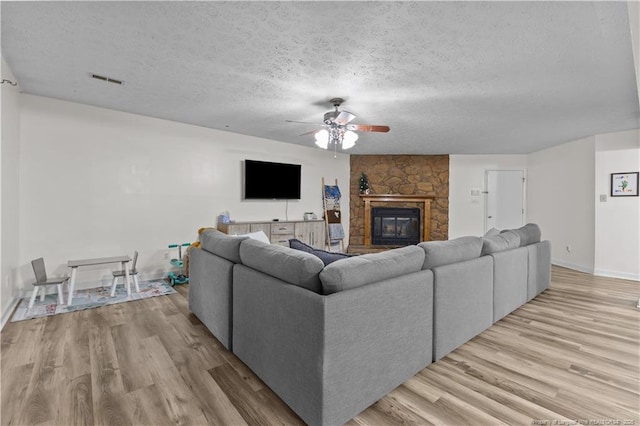 living area featuring visible vents, light wood-style flooring, a textured ceiling, a stone fireplace, and ceiling fan