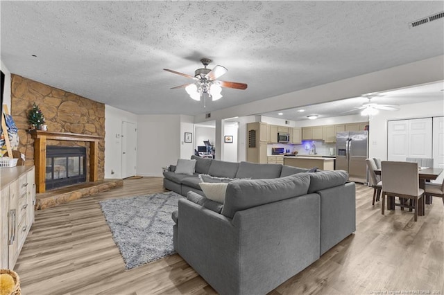living room with visible vents, a textured ceiling, a stone fireplace, light wood finished floors, and ceiling fan