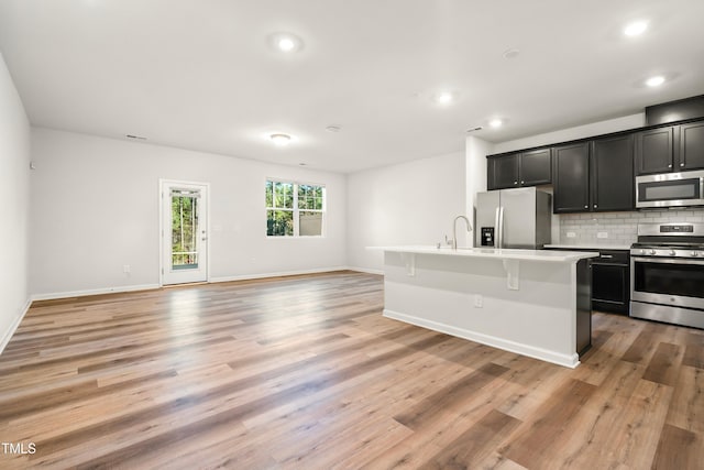 kitchen featuring dark cabinetry, a center island with sink, decorative backsplash, light wood-style floors, and appliances with stainless steel finishes