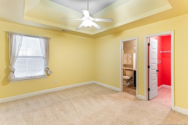 unfurnished bedroom featuring a walk in closet, baseboards, light colored carpet, a tray ceiling, and ensuite bathroom