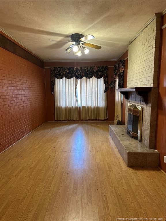 unfurnished living room with a textured ceiling, brick wall, a fireplace, light wood finished floors, and ceiling fan