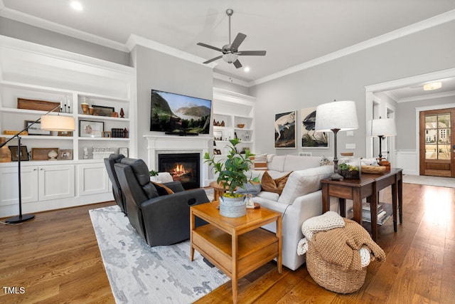 living area featuring built in shelves, ornamental molding, a glass covered fireplace, wood finished floors, and ceiling fan