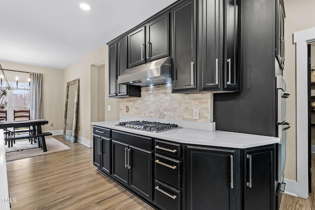 kitchen featuring under cabinet range hood, backsplash, light wood-style floors, dark cabinets, and stainless steel gas cooktop