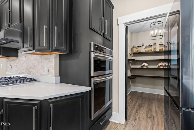 kitchen with under cabinet range hood, tasteful backsplash, wood finished floors, appliances with stainless steel finishes, and dark cabinets