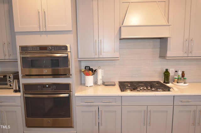 kitchen with custom exhaust hood, gray cabinetry, stainless steel appliances, light countertops, and backsplash