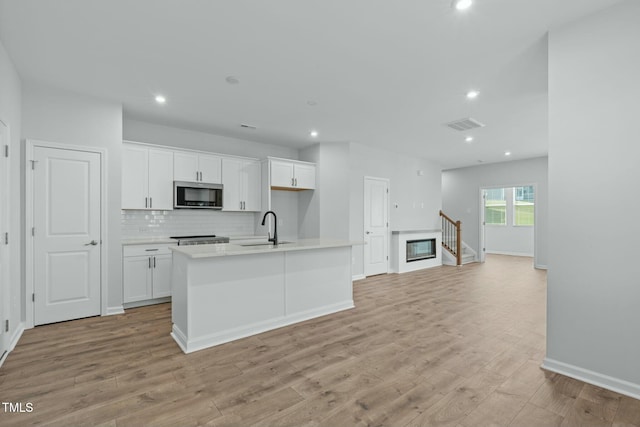 kitchen featuring stainless steel microwave, light wood-type flooring, an island with sink, white cabinetry, and a sink