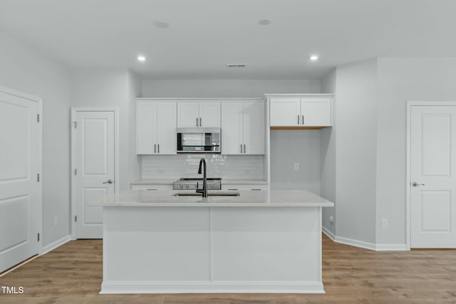 kitchen featuring a kitchen island with sink, stainless steel microwave, white cabinets, and light countertops