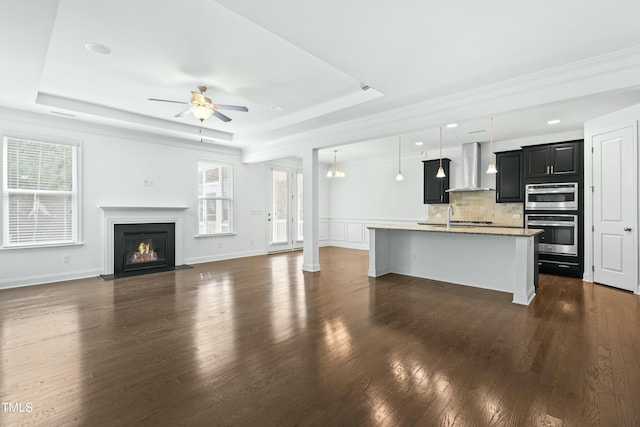 unfurnished living room with dark wood-style floors, baseboards, a tray ceiling, a fireplace with flush hearth, and ceiling fan