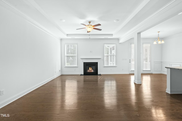 unfurnished living room featuring baseboards, a tray ceiling, a fireplace with flush hearth, dark wood-style flooring, and ceiling fan with notable chandelier