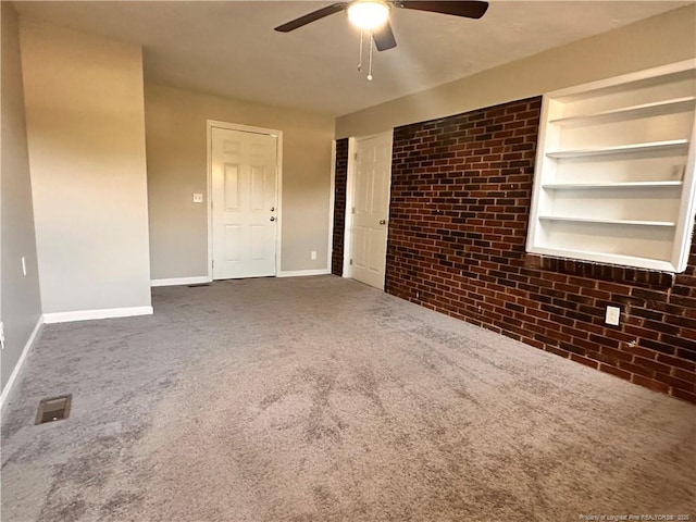 unfurnished living room featuring built in shelves, baseboards, brick wall, carpet floors, and ceiling fan