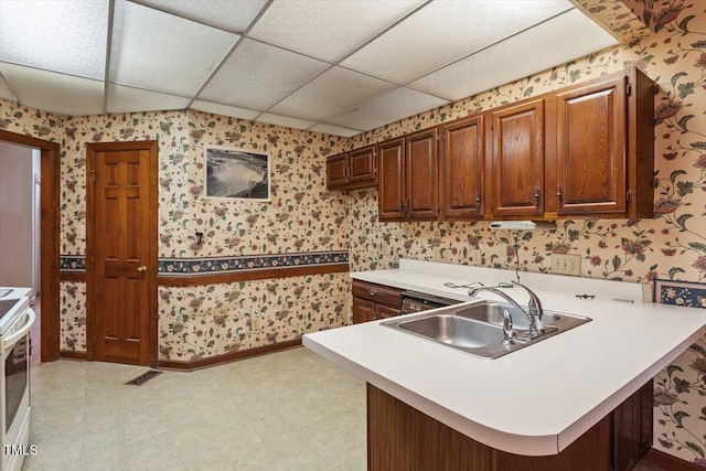 kitchen with wallpapered walls, a sink, a peninsula, light countertops, and a drop ceiling