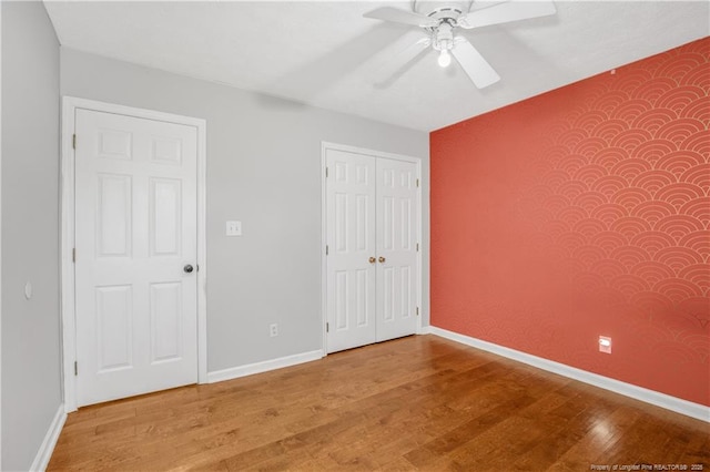 unfurnished bedroom featuring a ceiling fan, wood finished floors, a closet, baseboards, and an accent wall