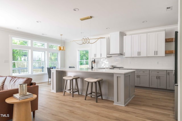 kitchen featuring backsplash, ornamental molding, light wood finished floors, a kitchen island with sink, and light countertops