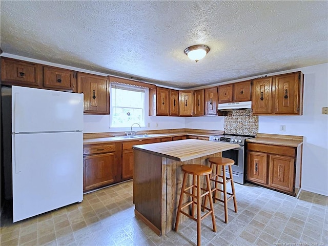 kitchen featuring under cabinet range hood, a sink, stainless steel range with electric cooktop, freestanding refrigerator, and brown cabinetry