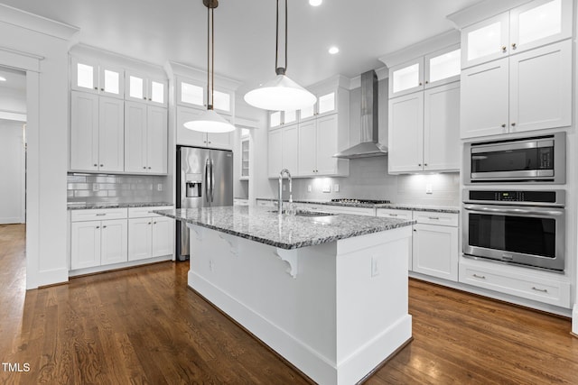 kitchen with wall chimney range hood, white cabinets, appliances with stainless steel finishes, a sink, and a center island with sink