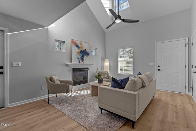 living room with high vaulted ceiling, baseboards, a ceiling fan, light wood-style floors, and a glass covered fireplace
