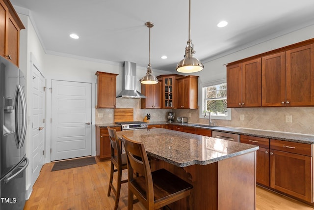 kitchen with appliances with stainless steel finishes, a sink, light wood finished floors, wall chimney exhaust hood, and a kitchen bar