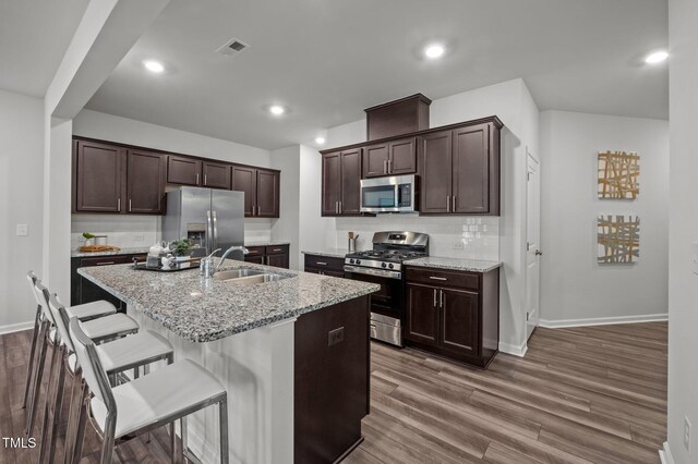 kitchen with appliances with stainless steel finishes, a sink, dark brown cabinetry, backsplash, and a center island with sink