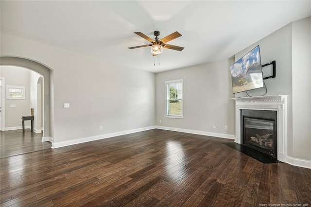 unfurnished living room with a ceiling fan, arched walkways, dark wood-type flooring, a fireplace with flush hearth, and baseboards