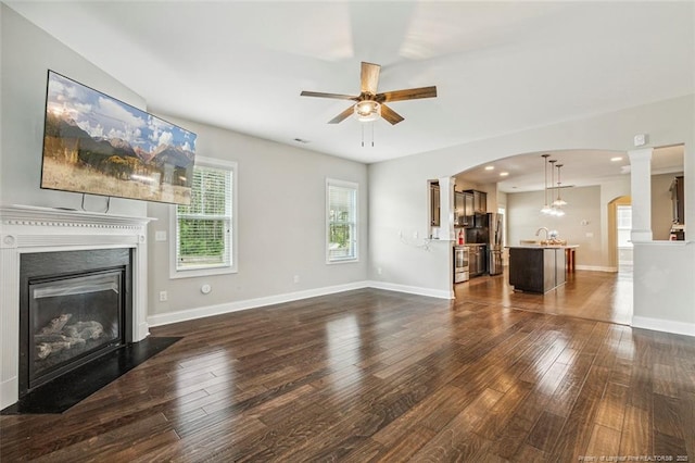 unfurnished living room with ceiling fan, dark wood-style floors, arched walkways, and a fireplace with flush hearth