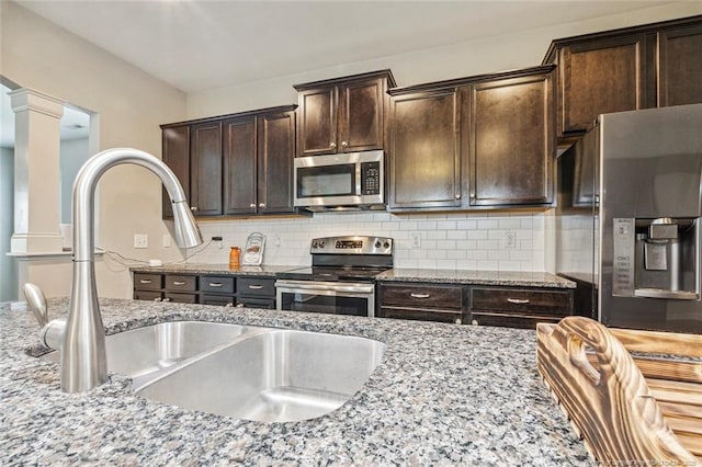 kitchen with appliances with stainless steel finishes, a sink, light stone counters, dark brown cabinets, and backsplash