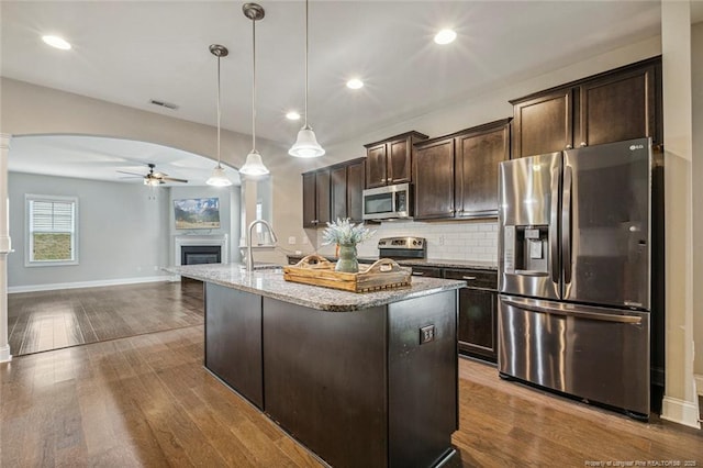 kitchen with stainless steel appliances, ceiling fan, ornate columns, dark brown cabinetry, and light stone counters
