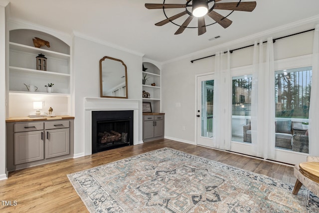 living room featuring ceiling fan, light wood-style flooring, a fireplace, ornamental molding, and built in features