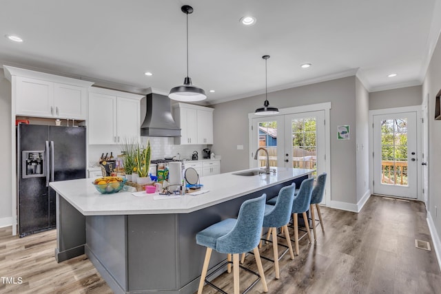 kitchen with wall chimney exhaust hood, a large island with sink, ornamental molding, black refrigerator with ice dispenser, and a sink
