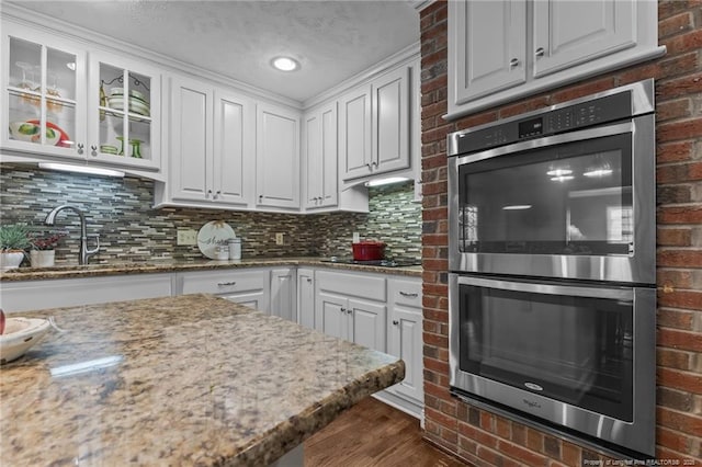 kitchen featuring glass insert cabinets, double oven, dark wood-type flooring, tasteful backsplash, and white cabinetry