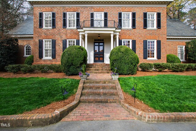 georgian-style home with a balcony, a shingled roof, a front lawn, and brick siding