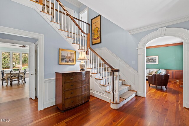 stairs with a ceiling fan, wood finished floors, ornamental molding, and wainscoting