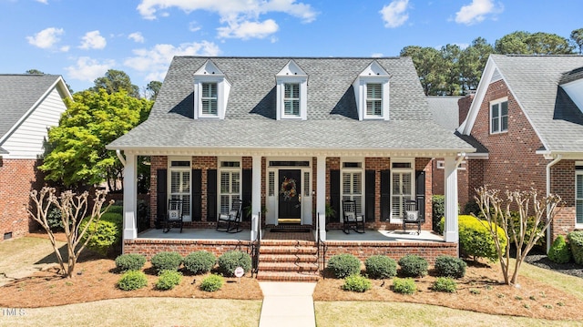 cape cod-style house featuring a shingled roof, a porch, and brick siding
