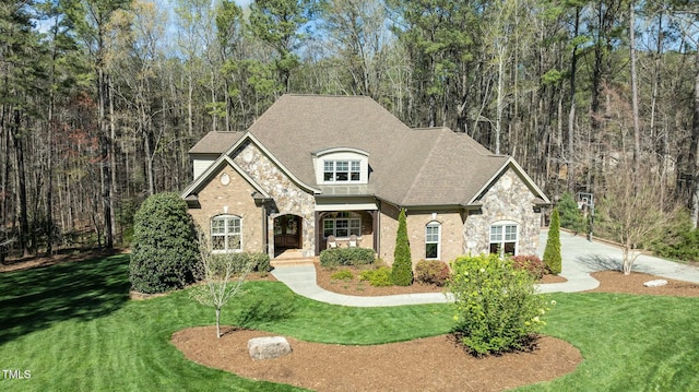 french country home featuring stone siding, a shingled roof, a view of trees, and a front lawn