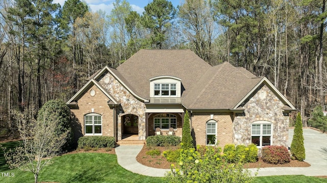 view of front of home with stone siding, covered porch, roof with shingles, and a front lawn