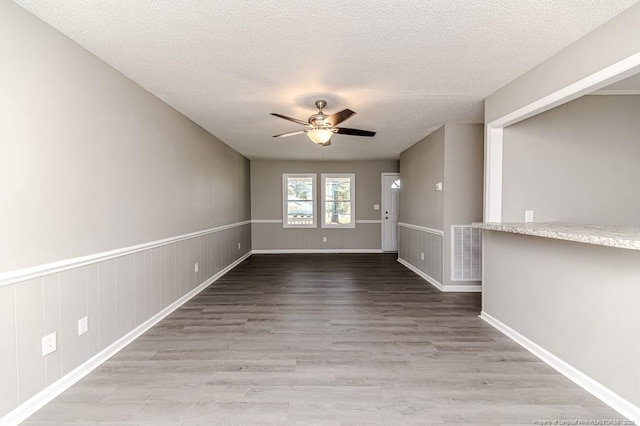 unfurnished room with a ceiling fan, a textured ceiling, visible vents, wood finished floors, and a wainscoted wall