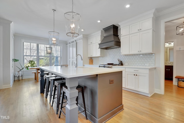 kitchen featuring decorative backsplash, a center island with sink, premium range hood, white cabinets, and a notable chandelier